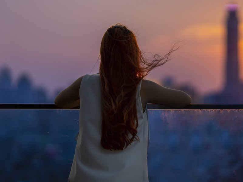 Woman looking and enjoying the sunset view from balcony with the sun setting behind skyscraper in busy urban downtown with loneliness for solitude, loneliness and dreaming of freedom lifestyle concept